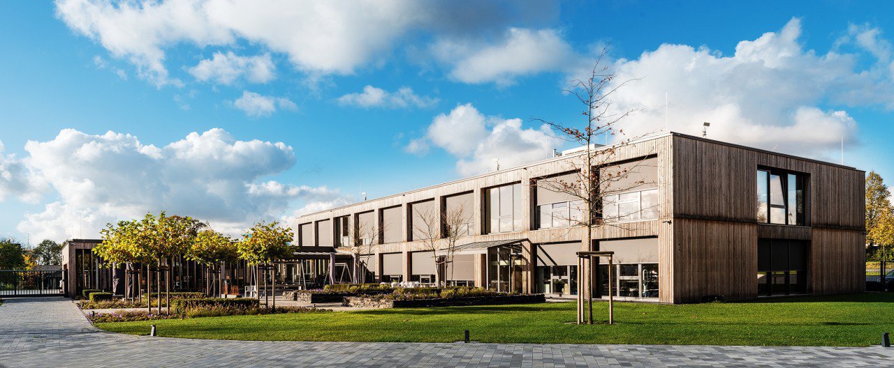 Moderne Holzarchitektur mit großen Fenstern, umgeben von Bäumen und Rasenfläche unter einem blauen Himmel.